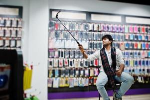 Indian man customer buyer at mobile phone store making selfie by smartphone on monopod stick. South asian peoples and technologies concept. Cellphone shop. photo