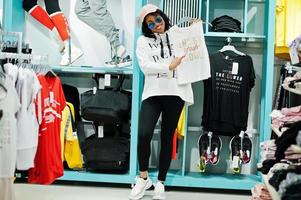 mujeres afroamericanas en chándales y gafas de sol comprando en un centro comercial de ropa deportiva contra estantes. tema de la tienda de deportes. ella sostiene la ropa superior. foto