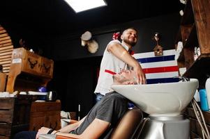Young bearded man washing head by hairdresser while sitting in chair at barbershop. Barber soul. photo