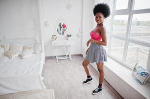 Attractive african american woman with afro hair wear on skirt pink top, posed at white room. Fashionable sexy black model. photo