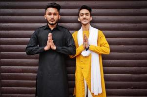 Two indian stylish mans friends in traditional clothes posed outdoor against wooden background and shows namaste hands sign. photo