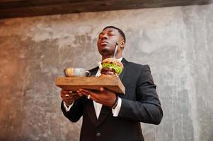 Respectable young african american man in black suit hold tray with double burger against gray wall. He breathes smell of a delicious hamburger.