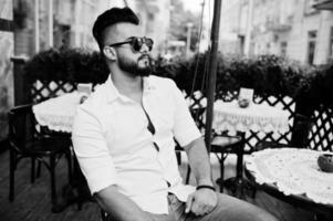 Stylish tall arabian man model in white shirt, jeans and sunglasses posed at street of city. Beard attractive arab guy sitting on outdoor cafe. photo