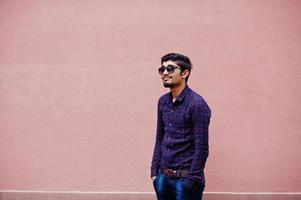 joven indio con camisa y gafas de sol posado contra la pared rosa. foto