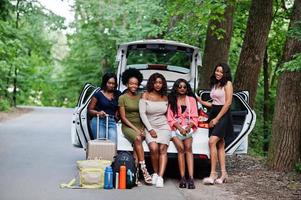 Group of five happy african american traveler girls sitting in car open trunk. photo