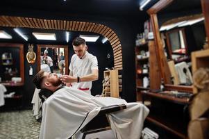 Young bearded man getting haircut by hairdresser while sitting in chair at barbershop. Barber soul. photo