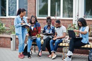 grupo de cinco estudiantes universitarios africanos que pasan tiempo juntos en el campus en el patio de la universidad. amigos afro negros que estudian en un banco con artículos escolares, computadoras portátiles. foto