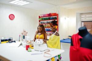 Two african dressmaker woman sews clothes on sewing machine and work with scissors at tailor office. Black seamstress girls. photo