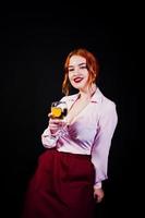 Gorgeous red haired girl in pink blouse and red skirt with glass of wine at hand isolated on black. photo