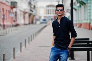 elegante modelo indio con ropa informal, camisa negra y gafas de sol posadas al aire libre en la calle de la india. foto