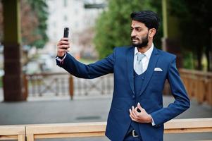 hombre de negocios indio de barba elegante con bindi en la frente, vestido con traje azul posado al aire libre y haciendo selfie en el teléfono móvil. foto