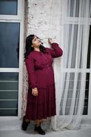 Attractive south asian woman in deep red gown dress posed at studio against windows in night. photo