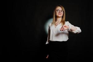 Studio portrait of blonde businesswoman in glasses, white blouse and black skirt holding laptop and looking at watches against dark background. photo