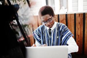 African man in traditional clothes and glasses sitting behind laptop at outdoor caffe. photo