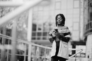 atractiva mujer afroamericana con rastas en chaqueta posada cerca de barandillas contra un moderno edificio de varios pisos con teléfono móvil. foto