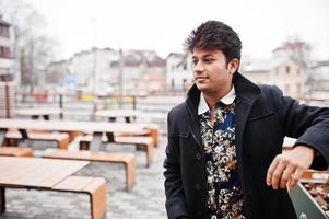 Attractive and handsome stylish indian man wear on black coat standing in the terrace of modern lounge cafe at his work break. photo