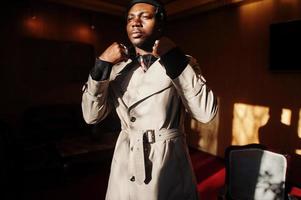 Handsome african american man posing  inside room with sunlight shadows in black hat and beige coat. photo