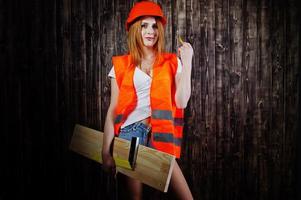 Engineer woman in orange protect helmet and building jacket against wooden background holding board and ruler. photo