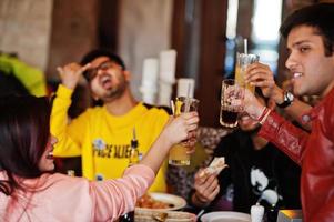 grupo de amigos asiáticos comiendo pizza y animando durante la fiesta en la pizzería. gente india feliz divirtiéndose juntos, comiendo comida italiana y sentados en el sofá. foto