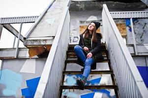 ropa de niña con sudadera verde larga, jeans y sombreros negros en un lugar industrial. foto