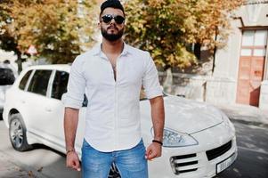 Elegante modelo de hombre árabe alto con camisa blanca, jeans y gafas de sol en la calle de la ciudad. chico árabe rico y atractivo con barba contra un auto todoterreno blanco. foto