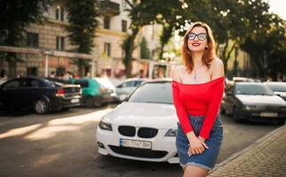 Attractive redhaired woman in eyeglasses, wear on red blouse and jeans skirt posing at street against white sport car. photo