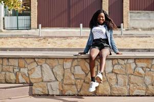 Modelo delgado afroamericano de piel oscura posado en pantalones cortos negros y chaqueta de jeans con teléfono móvil. foto