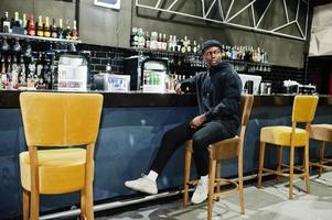 Handsome african american man posing  inside night club in black hat, sitting on bar counter. photo