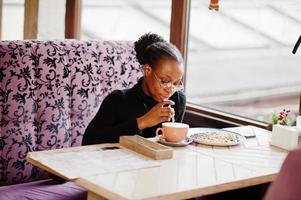 African woman in black sweater and eyeglasses posed at cafe, sitting by table with dessert and cup of coffee. photo