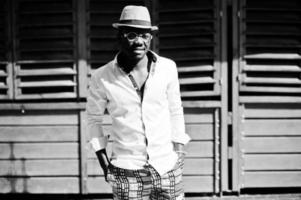 elegante hombre afroamericano con camisa blanca y pantalones de colores con sombrero y gafas al aire libre. chico modelo de moda negro. foto
