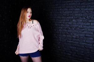 Studio shot of brunette girl at pink sweater with jeans shorts against black brick wall. photo