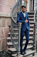 hombre indio de barba elegante con bindi en la frente, vestido con traje azul posado al aire libre contra escaleras de hierro y mostrando el pulgar hacia arriba. foto