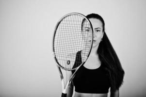 Black and white portrait of beautiful young woman player in sports clothes holding tennis racket while standing against white background. photo