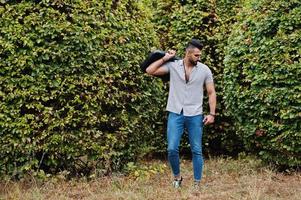 Fashionable tall arab beard man wear on shirt, jeans and sunglasses posed on park and hold bag case. photo