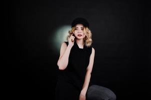 Studio portrait of blonde girl in black wear and cap with mobile phone at hand against dark background. photo