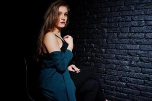 hermosa chica morena vestida con una chaqueta negra y verde, sentada y posando en una silla en el estudio contra una pared de ladrillo oscuro. retrato de modelo de estudio. foto