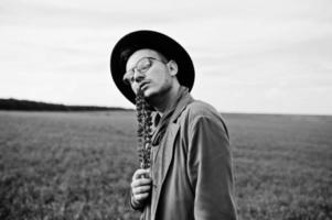 Stylish man in glasses, brown jacket and hat posed on green field. photo