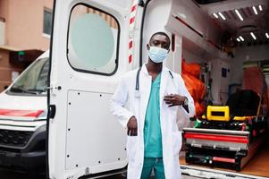 African male paramedic in face protective medical mask standing in front of ambulance car. photo