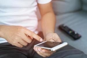 young man using television remote control and mobile smart phone  is sitting on a sofa. on or off tv.  Leave space to write a description of the message. photo