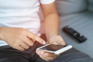 un joven que usa el control remoto de la televisión y un teléfono inteligente móvil está sentado en un sofá. encendido o apagado de la televisión. deje espacio para escribir una descripción del mensaje. foto