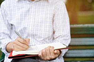 cerrar mano joven está sentado en una silla de mármol. usando el bloc de notas de la conferencia del registro de escritura de la pluma en el libro en el parque público. foto