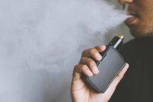 photo portrait young man with beard hold and smoking his electronic cigarette blowing a stream of smoke spread. dark black, grey background