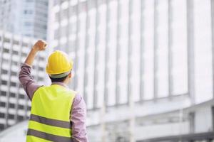 Close up backside view of engineering male construction worker stand holding safety yellow helmet and wear reflective clothing for the safety of the work operation. outdoor of building background. photo