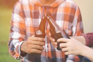 viajando tres jóvenes amigos que se divierten juntos, se relajan en el campamento de senderismo y beben botellas de cerveza y disfrutan. concepto de aventura de viaje de camping de vacaciones. fondo de vista de río de bosque de hierba. foto