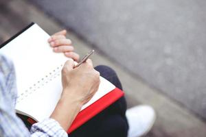 close up hand young man are using pen writing Record Lecture note pad into the book sitting on the chair outdoor. Blank book page copy  write a message. photo