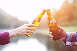 viajando dos jóvenes amigos que se divierten juntos, se relajan en el campamento de senderismo y beben botellas de cerveza y disfrutan. concepto de aventura de viajes de vacaciones. fondo de vista de río de bosque de montaña. foto