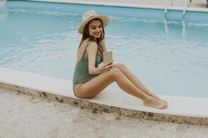 Young woman sitting by the swimming pool and taking selfie photo with  mobile phone in the house backyard