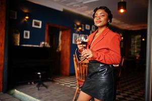 Portrait of african american woman, retro hairstyle with eyeglasses, wear orange jacket and leather skirt posing at restaurant with glass of wine. photo