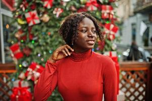 retrato de una mujer africana de pelo rizado con cuello de tortuga rojo de moda posando contra decoraciones navideñas, tema de la víspera de año nuevo. foto