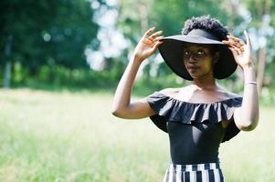 Portrait of gorgeous african american woman 20s in wear in black and white stripe pants and summer hat posing at green grass in park. photo